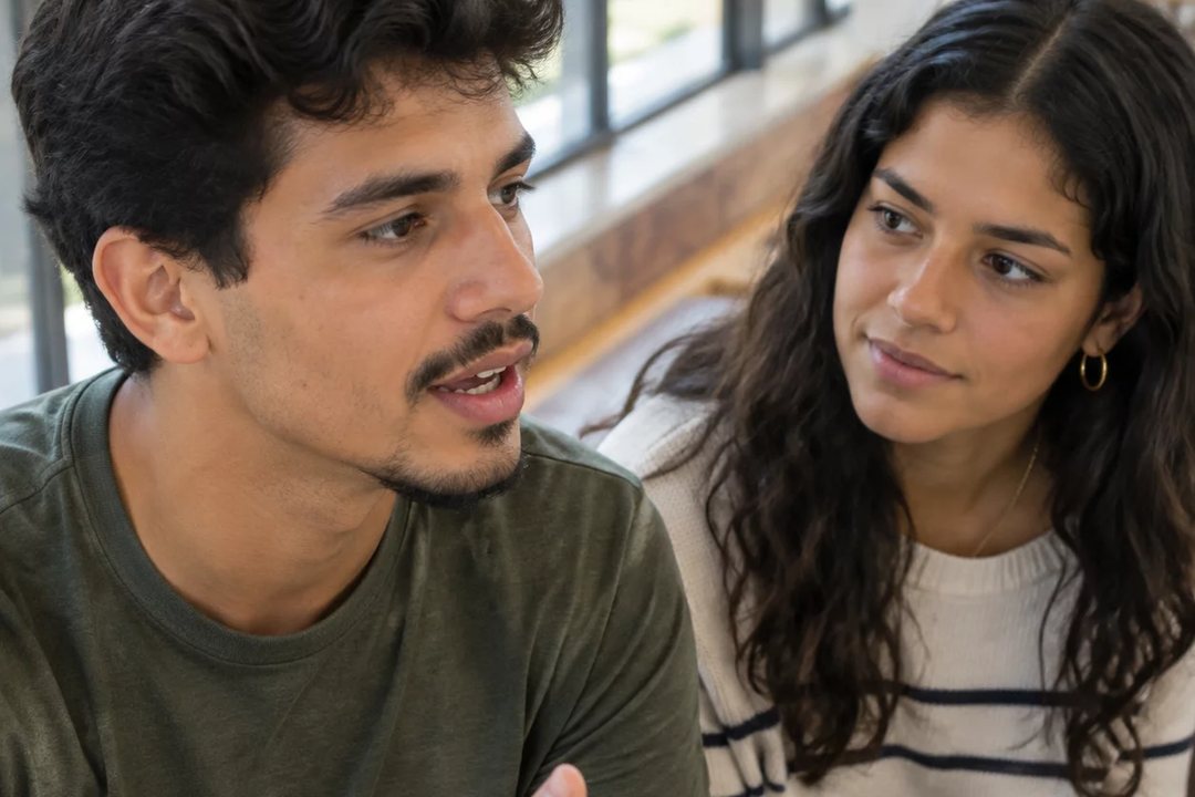 Três participantes conversam ao redor de uma mesa comprida numa sala clara de janelas altas